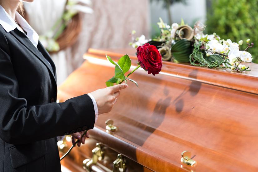 Frau in schwarzem Anzug hält eine rote Rose über einen Holzsarg, der mit Blumen dekoriert ist.