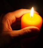 Eine Hand hält eine brennende orangefarbene Kerze in einem dunklen Umfeld.