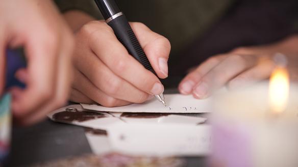 Nahaufnahme einer Hand, die mit einem Kugelschreiber auf einem Zettel schreibt, im Hintergrund eine brennende Kerze.